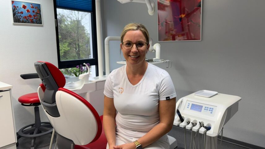 Dr. Daniela Meusch sitzt mit weißem T-Shirt in einem Behandlungsstuhl in einer Zahnarztpraxis mit rotem Stuhl und zahnärztlichen Geräten