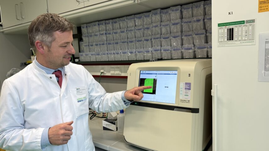 Professor Michael Wolf von der RWTH Aachen steht im molekularbiologischen Forschungslabor und zeigt auf den Bildschirm eines Laborgeräts mit einer grünen Grafik, er trägt einen weißen Laborkittel und steht vor einem Regal mit vielen kleinen Behältern.